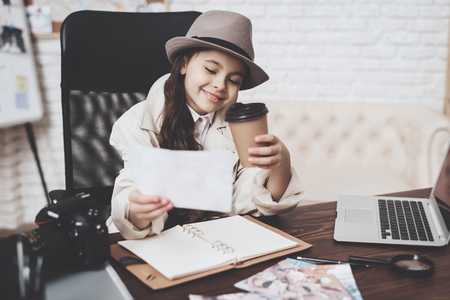Private detective agency. Little girl in cloak and hat is sitting at desk looking at different photos and drinking coffee.の写真素材