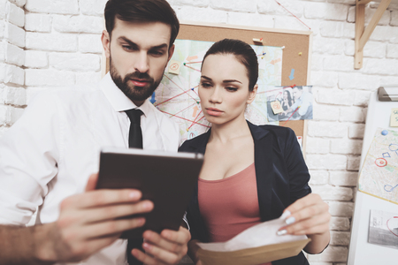 Private detective agency. Man in tie and woman in jacket are looking at clues on tablet in office.の写真素材