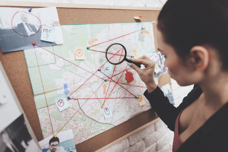 Private detective agency. Woman in jacket is looking at clue map with magnifying glass in office.