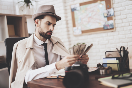 Private detective agency. Man in hat and cloak is sitting at table counting money.の写真素材