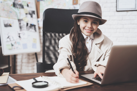 Private detective agency. Little girl in cloak and hat is sitting at desk taking notes near laptop.の写真素材