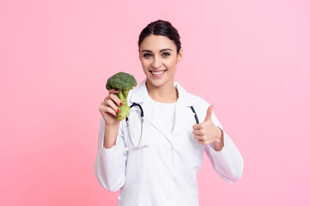 Portrait of smiling female doctor in white gown with stethoscope holding broccoli isolated.の写真素材
