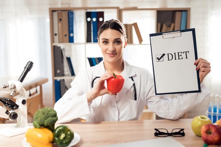 Female doctor in white gown sitting at desk in office with microscope and stethoscope. Woman is holding diet sign and pepper.の写真素材