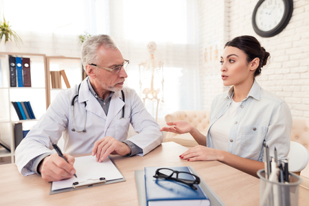 Doctor in white gown with stethoscope and female patient in office. Patient is telling symptoms.の写真素材