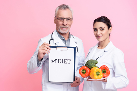 Portrait of smiling male and female doctors in white gowns with stethoscopes holding diet sign and plate of vegetables isolated.の写真素材