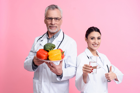 Portrait of smiling male and female doctors in white gowns with stethoscopes holding water and plate of vegetables isolated.の写真素材