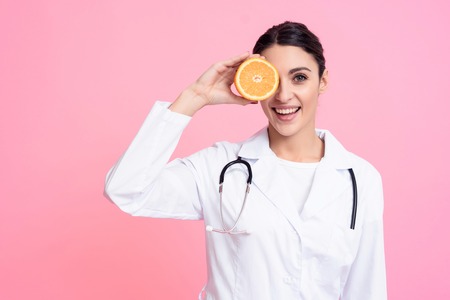 Portrait of smiling female doctor in white gown with stethoscope holding orange isolated.の写真素材