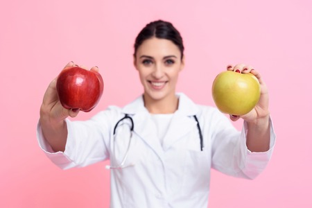 Portrait of smiling female doctor in white gown with stethoscope holding red and yellow apples isolated.の写真素材