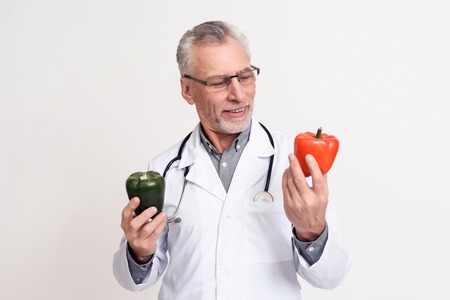 Portrait of doctor in white gown with stethoscope holding red and green peppers isolated.の写真素材