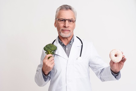 Portrait of doctor in white gown with stethoscope holding broccoli and donut isolated.の写真素材