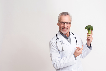 Portrait of smiling doctor in white gown with stethoscope holding broccoli isolated.の写真素材