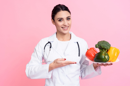 Portrait of smiling female doctor in white gown with stethoscope holding plate of vegetables isolated.の写真素材