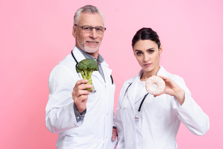 Portrait of male and female doctors in white gowns with stethoscopes holding broccoli and donut isolated.の写真素材