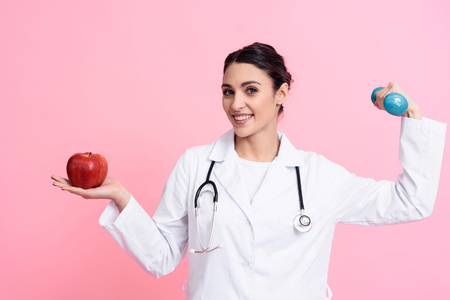 Portrait of smiling female doctor in white gown with stethoscope holding red apple and dumbbell isolated.の写真素材