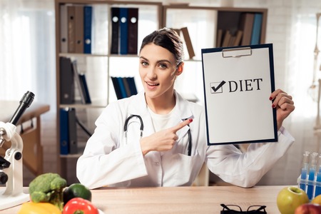 Female doctor in white gown sitting at desk in office with microscope and stethoscope. Woman is holding diet sign.の写真素材