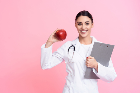 Portrait of smiling female doctor in white gown with stethoscope holding red apple isolated.の写真素材