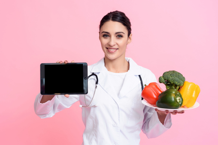 Portrait of smiling female doctor in white gown with stethoscope holding plate of vegetables and tablet isolated.の写真素材