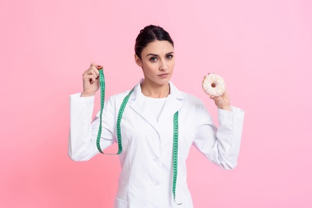 Portrait of female doctor in white gown with stethoscope holding measuring tape and donut isolated.の写真素材