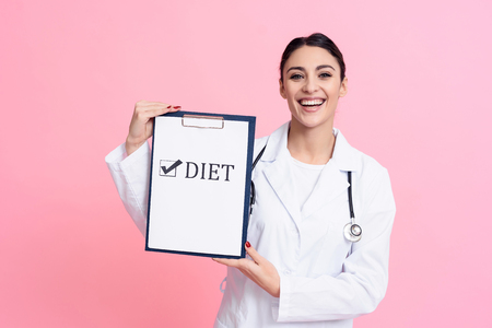 Portrait of smiling female doctor in white gown with stethoscope holding diet sign on clipboard isolated.の写真素材