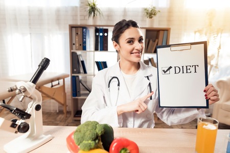 Female doctor in white gown sitting at desk in office with microscope and stethoscope. Woman is holding diet sign.の写真素材