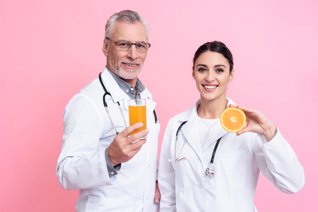 Portrait of smiling male and female doctors in white gowns with stethoscopes holding juice and orange isolated.の写真素材
