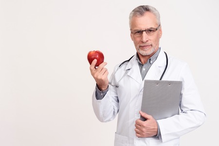 Portrait in white gown of serious doctor with stethoscope and clipboard holding apple isolated.の写真素材