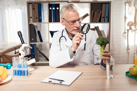 Doctor in white robe sitting at desk in office. Man is looking at broccoli with magnifying glass.の写真素材