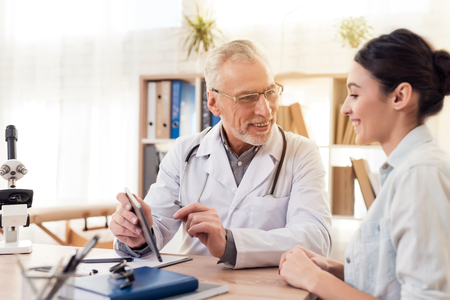 Doctor in white gown with stethoscope and female patient in office. Doctor is using tablet.の写真素材