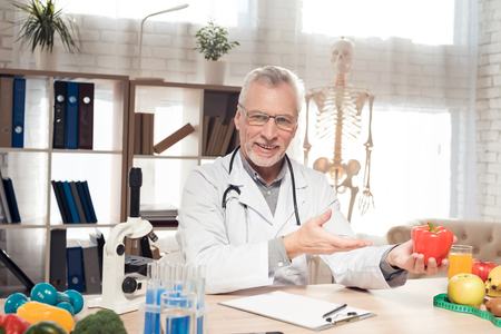 Doctor in white robe sitting at desk in office with microscope and stethoscope. Man is holding red pepper.の写真素材