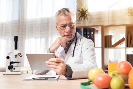 Doctor in white robe sitting at desk in office with microscope and stethoscope. Man is holding tablet.の写真素材