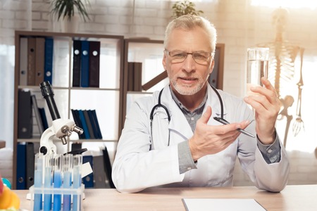 Doctor in white robe sitting at desk in office with microscope and stethoscope. Man is holding water.の写真素材