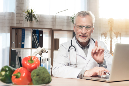 Doctor in white robe sitting at desk in office with microscope and stethoscope. Man is working on laptop.の写真素材