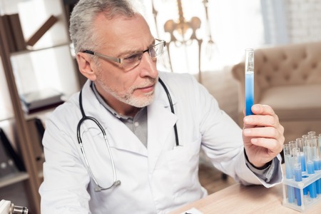 Doctor in white robe sitting at desk in office with microscope and stethoscope. Man is holding medicine glass.の写真素材