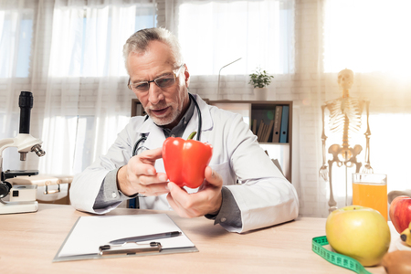 Doctor in white robe sitting at desk in office with microscope and stethoscope. Man is holding red pepper.の写真素材