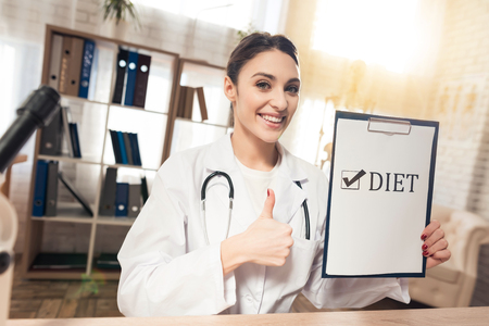 Female doctor in white gown sitting at desk in office with microscope and stethoscope. Woman is holding diet sign.の写真素材