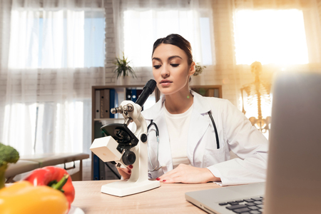 Female doctor in white gown sitting at desk in office with microscope and stethoscope. Woman is looking in microscope.の写真素材