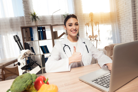 Female doctor in white gown sitting at desk in office with microscope and stethoscope. Woman is giving thumbs up.の写真素材