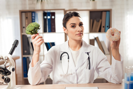 Female doctor in white gown sitting at desk in office with microscope and stethoscope. Woman is holding broccoli and donut.の写真素材
