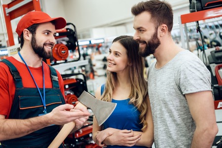 Salesman in red shirt and baseball cap is showing couple of clients new axe in power tools store.の写真素材