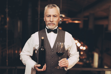 Experienced sommelier in bow tie is standing with bottle of wine and wine glass. Wine tasting. Professional degustation expert in winemaking.の写真素材