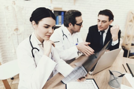 Young nurse examines x-ray at medical office. Blurred doctor and patient in background. Adult man in business suit is being examined in clinic.の写真素材