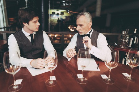Confident young and old sommeliers discuss quality of wine drink in restaurant. Glasses on table. Confident sommelier makes wine list in restaurant.の写真素材