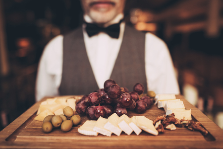 Close up. Sommelier is holding snacks for red wine on wooden plate. Grapes. Cheese. Olives. Walnut. Waiter with wooden tray.の写真素材