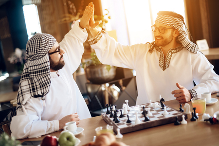 Two arab businessmen in thawb high five behind chessboard at hotel room.の写真素材
