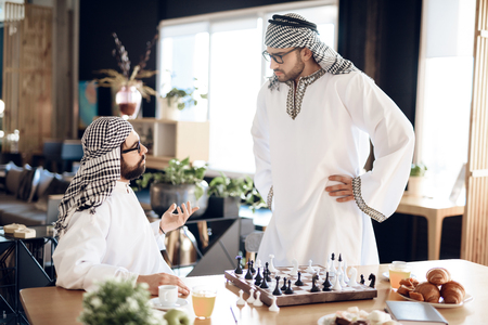 Two arab businessmen in thawb arguing playing chess at table at hotel room.の写真素材