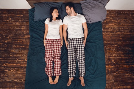 Top view. Handsome man and beautiful woman in pajama lie straight on bed and look at each other. Young couple of people are at domestic atmosphere.の写真素材