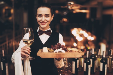 Joyful elegant waitress holding glass of white wine, plate of cheese and fruit, standing near bar. Wine tasting.の写真素材