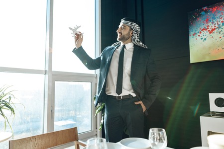 Bearded arab businessman in suit standing at window with model airplane.の写真素材