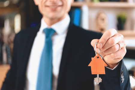 Realtor in suit sitting at desk in office. Realtor is showing keys with cutout icon of new house.の写真素材