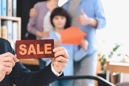 Realtor in suit sitting at desk in office. Realtor is presenting sale sign with family in background.の写真素材